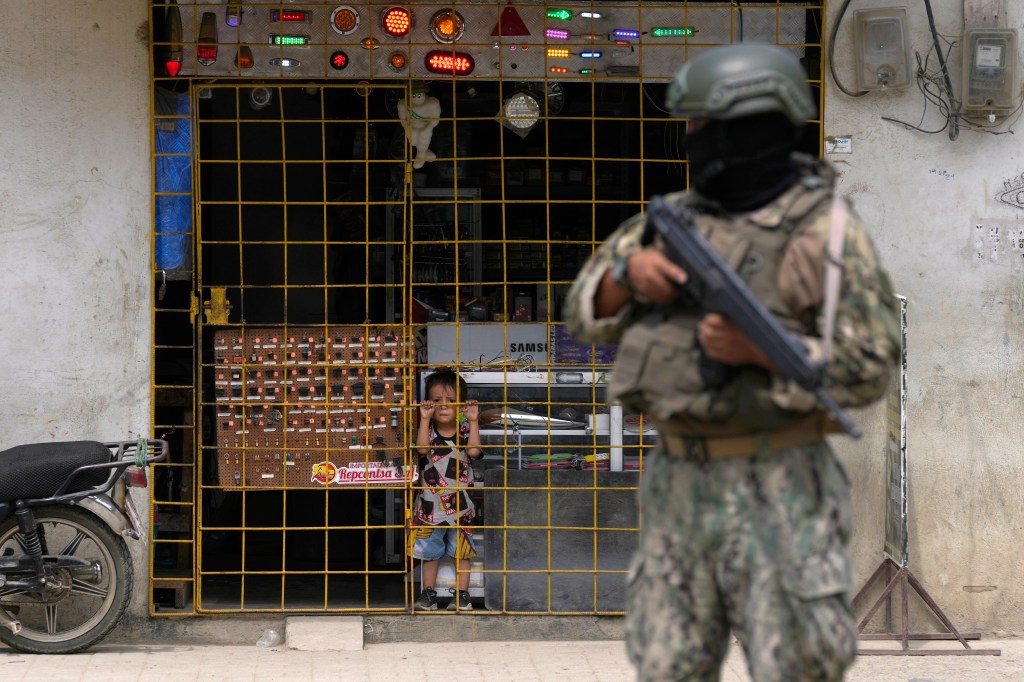 A boy watches from behind the bars of an open electronics store as a soldier stands guard at a security check point placed by the army in Duran, across a bridge from Guayaquil, Ecuador, Monday, Aug. 14, 2023.