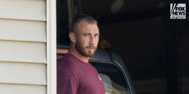 Richard Tobin in blue shorts and and a purple t-shirt looks suspicious as he peeks from the garage