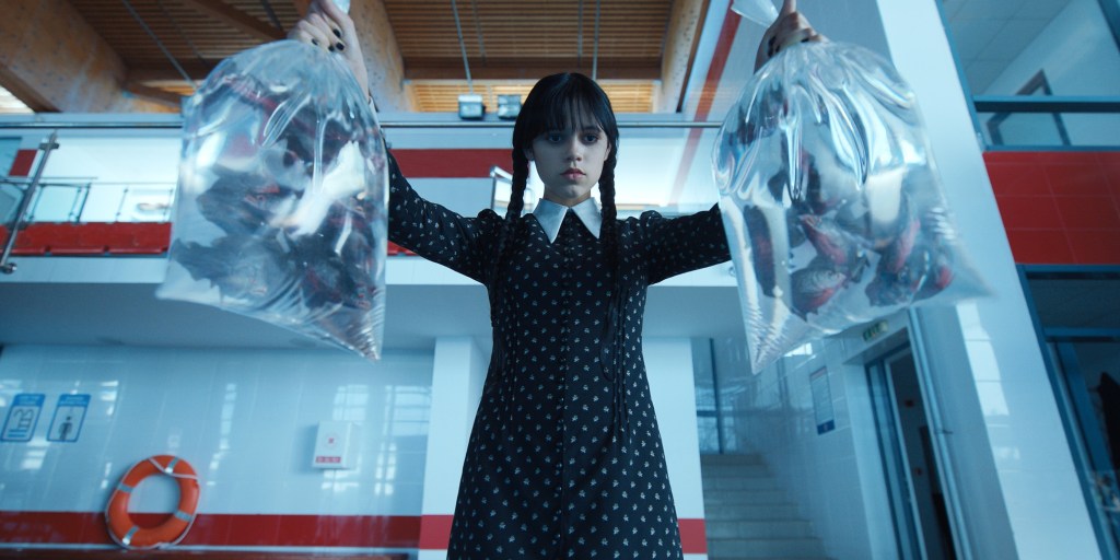 Wednesday Addams, portrayed by Jenna Ortega, holding a bag of piranhas in each hand over a pool. 
