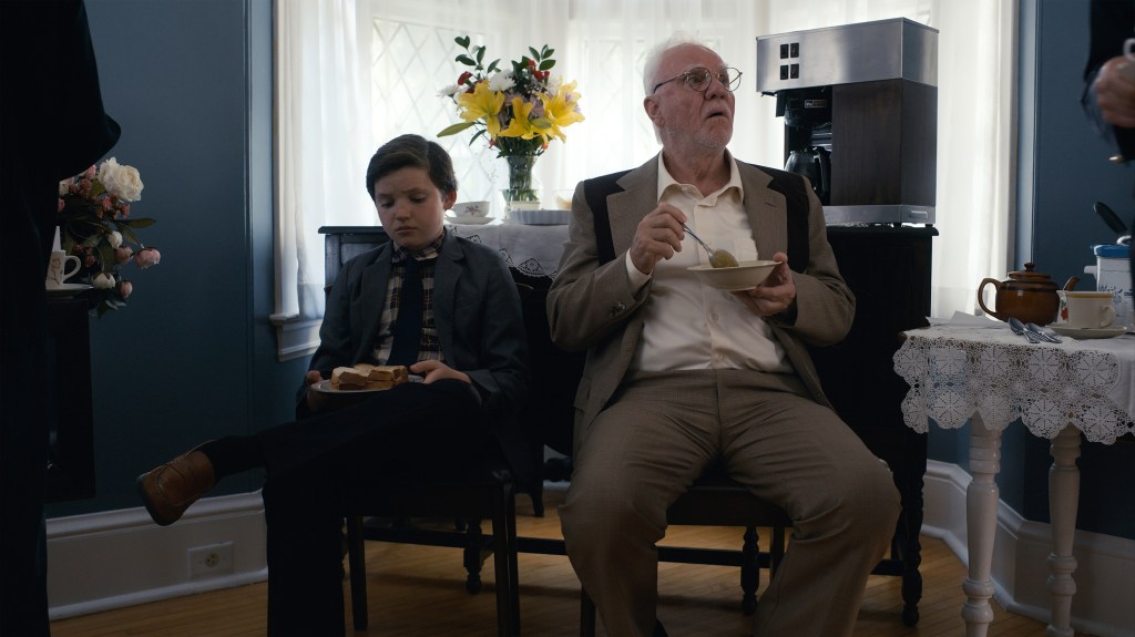 Mark (Benjamin Evan Ainsworth) and his grandfather, Pop Critch (Malcolm McDowell) try to enjoy a snack. They're seated side by side on wooden chairs and they both look disappointed. Mark is holding a sandwich and Pop a bowl of soup.