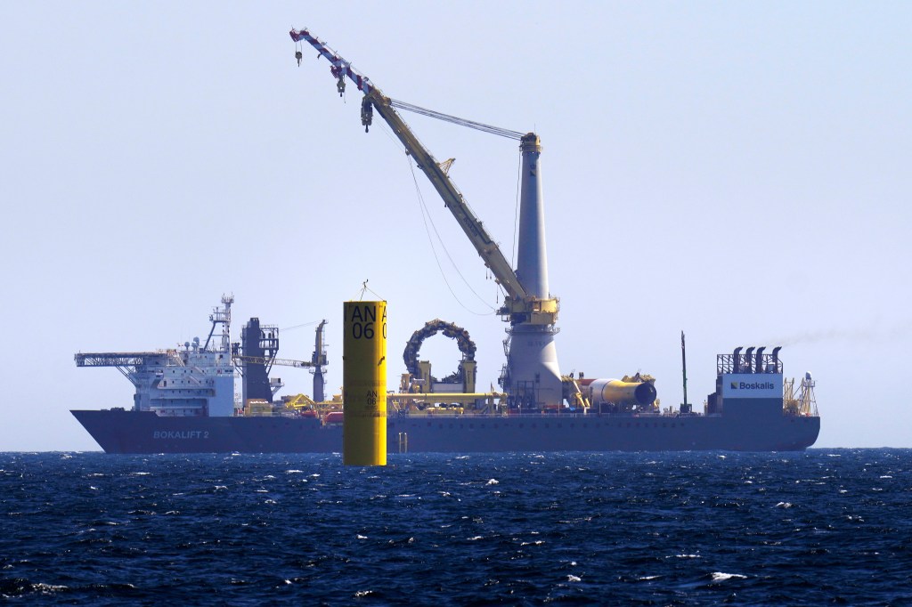 A monopile, center, looms above the waterline prior to the installation of a tower, blades and turbine sections, as the crane ship "Bokalift 2" motors through the South Fork Wind project, Tuesday, July 11, 2023