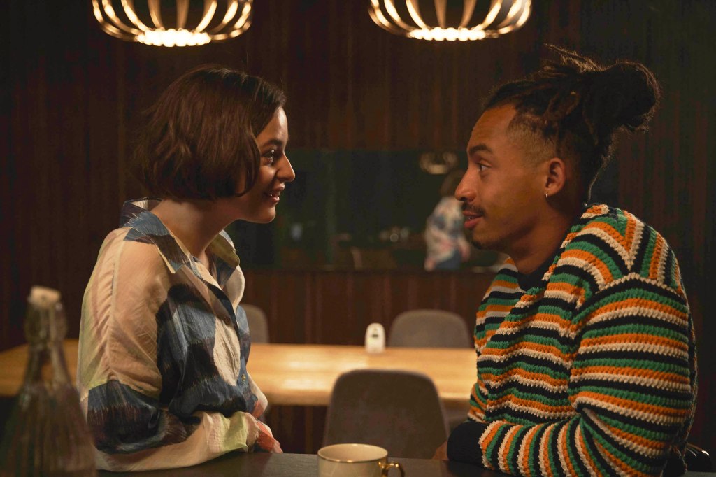 Céline Buckens and Jordan Stephens as Tasha and Sam. They're in what appears to be a bar and are facing each other and smiling at each other. There's a coffee cup between them.