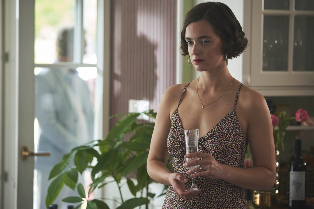 Céline Buckens as Tasha. She's wearing a cocktail dress and looks forlorn as she holds a glass of champagne and pouts.