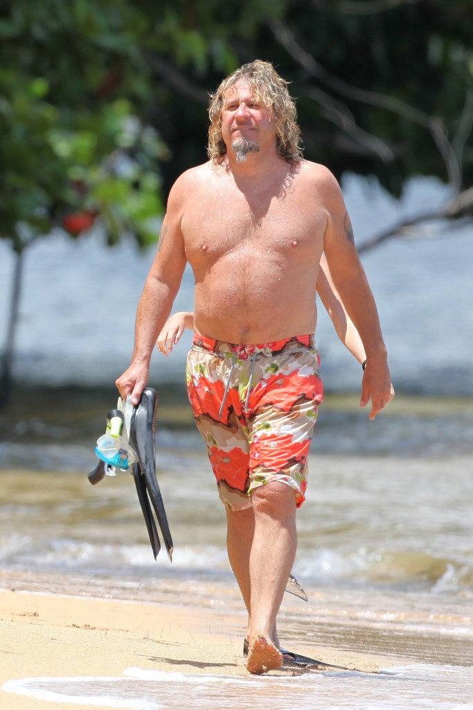 Sammy Hagar walking on the beach in shorts and no shirt, holding snorkel and flippers. 