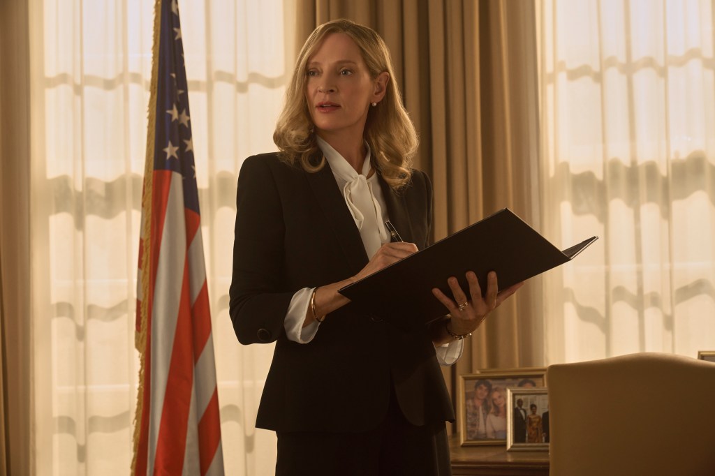 Uma Thurman standing by an American flag holding a binder. 