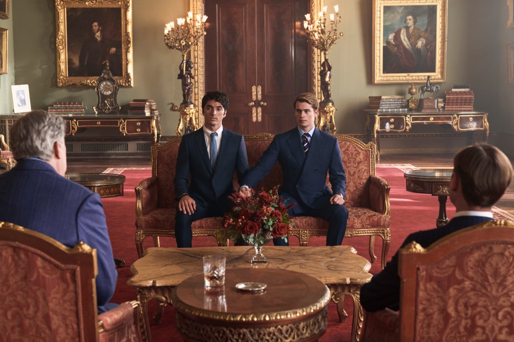 Alex (Taylor Zakhar Perez) and Prince Henry (Nicholas Galitzine) hold hands sitting next to each other on a couch. 