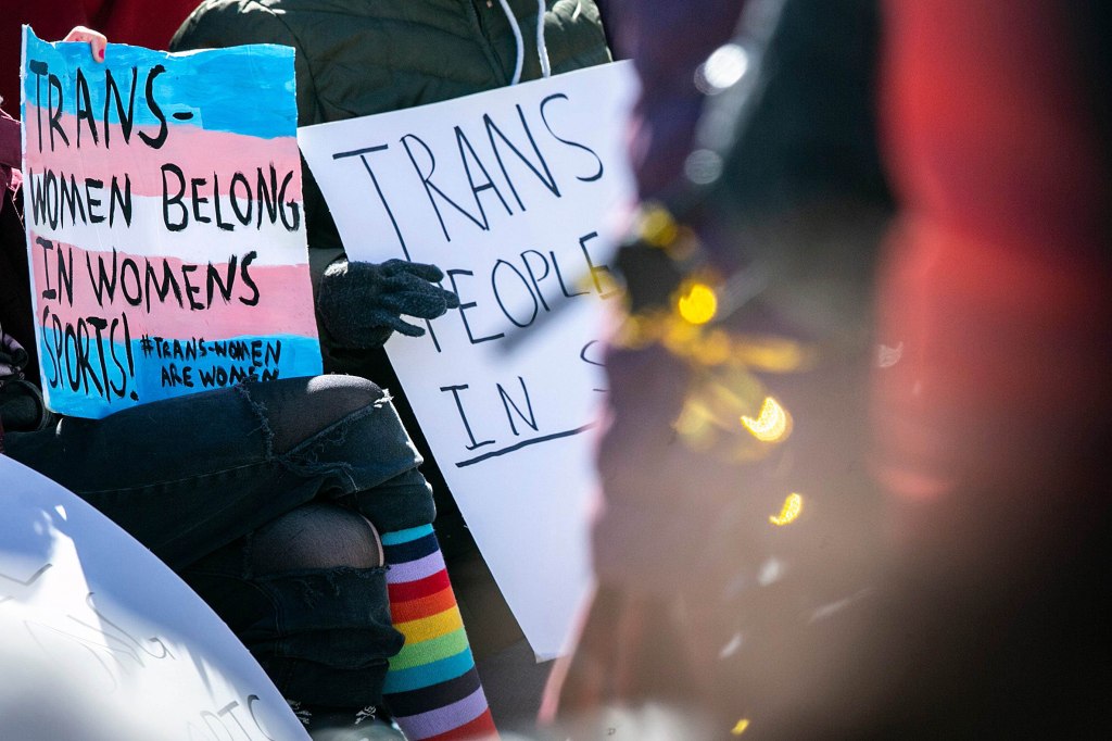 People hold signs reading "Trans women belong in womens sports! #TransWomenAreWomen" during a protest against House File 2416, a bill signed by Gov. Kim Reynolds which immediately prohibited transgender women and girls from competing in female sports offered by Iowa schools, colleges and universities, Friday, March 11, 2022, on the Pentacrest in Iowa City, Iowa.