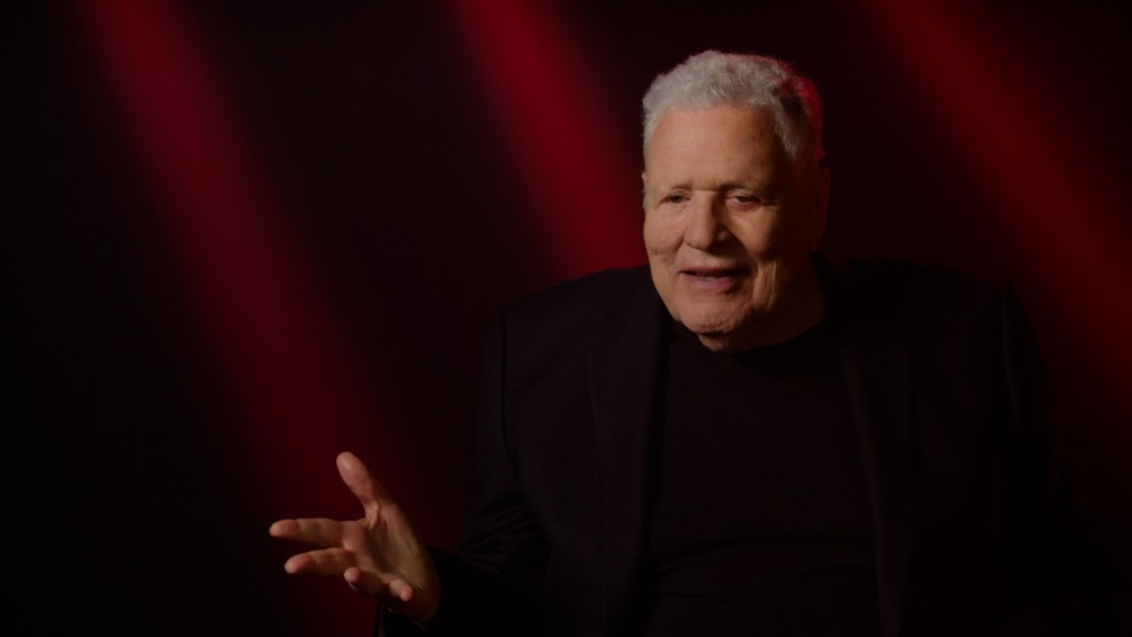 Steve Binder in a recent photograph taken from the Paramount+ documentary about the Elvis comeback special. He's got grey hair, is wearing all-black and is gesticulating with his right hand as if he's making a point.