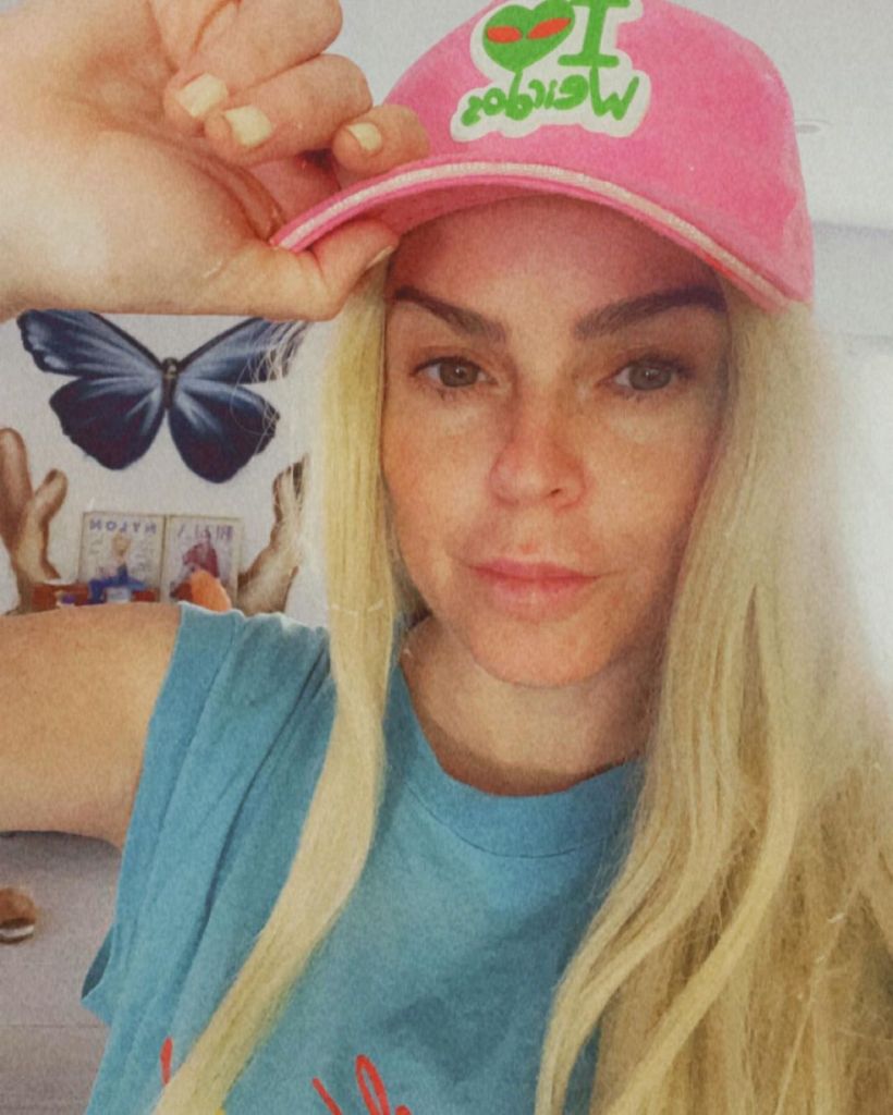 Taryn Manning looking at the camera with blonde hair and a pink hat. 