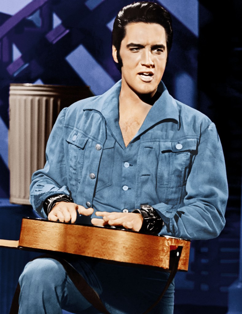 Elvis Presley in a color photo during the NBC special. He's wearing all-blue and is tapping out a rhythm on an acoustic guitar that he's holding on one knee.