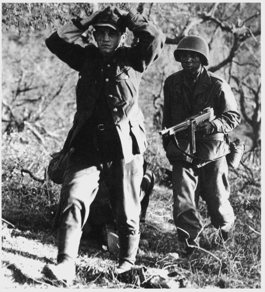 A 761st Tank Battalion soldier captures a German soldier in France. The soldier is carrying a Thompson machine gun which he has trained on the German soldier, whose arms are behind his head in surrender.