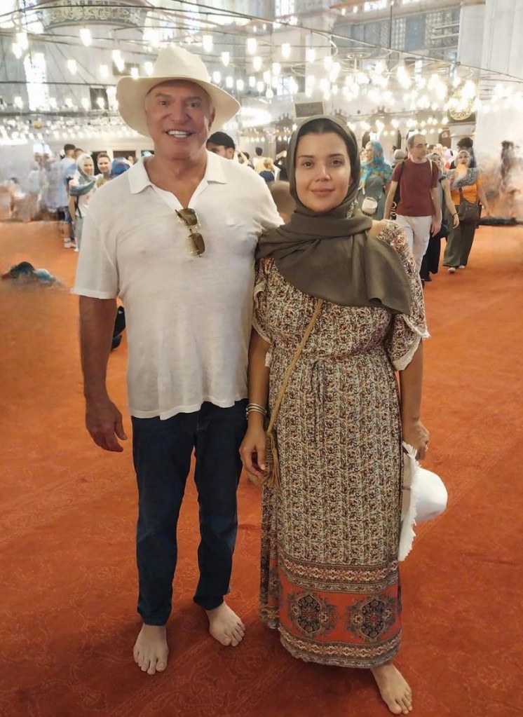 Man and woman standing in front of mosque. Man in jeans and white shirt and wide brim hat, woman with scarf over head and floral dress holding hat.