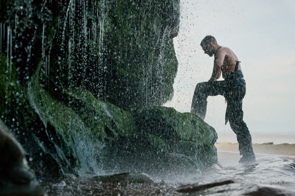 King Arthur (Ian De Caestecker) with his leg up on a rock washing himself in a waterfall. 
