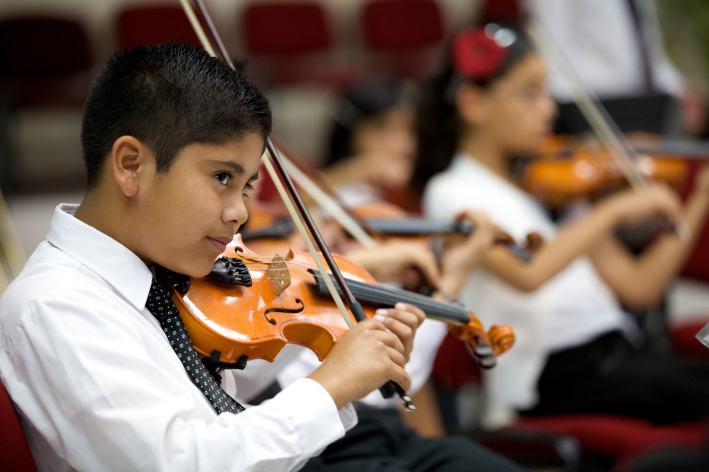 Young violinists play in open concert with the orchestra. Focus on foreground.