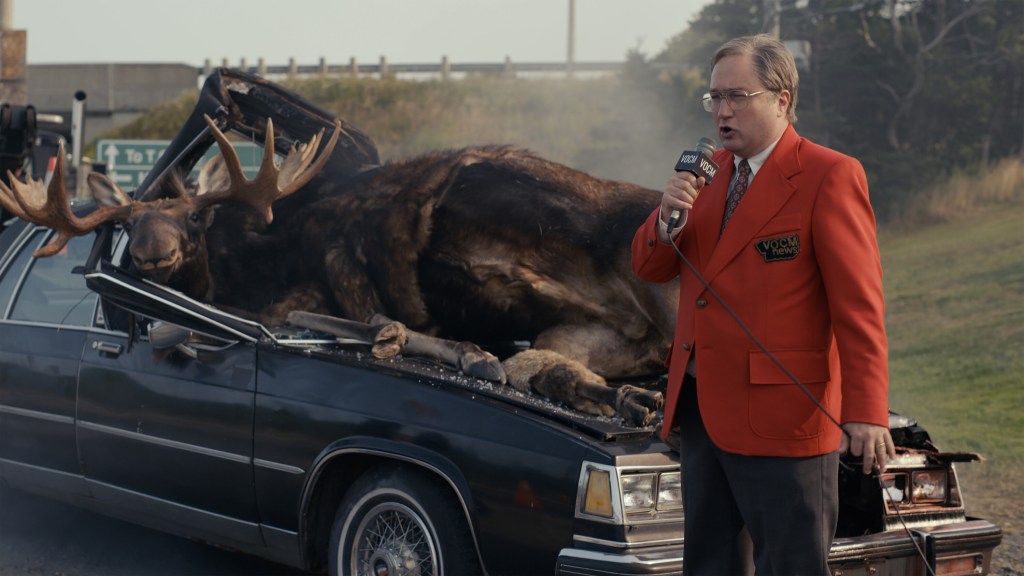 Mark's dad, Mike (Mark Critch) delivers a radio report on a car/moose accident in front of the Critch house, which is next-door to the local radio station. He's wearing a red blazer and is holding a microphone. He's standing in front of a smashed-up car with two dead moose lying where the windshield used to be.