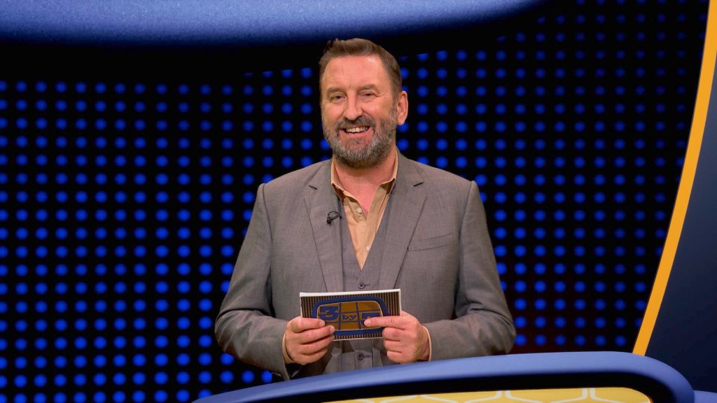 "3X3" host Lee Mack. He's smiling on the set of the show and is holding a card in his hand that says "3X3" on it. He's wearing a grey suit and a brownish shirt without a tie.