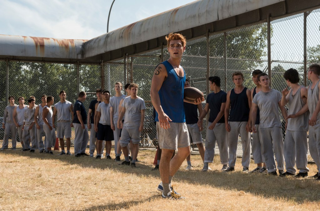 Archie (KJ Apa) holding a football standing in a yard in front of a crowd. 
