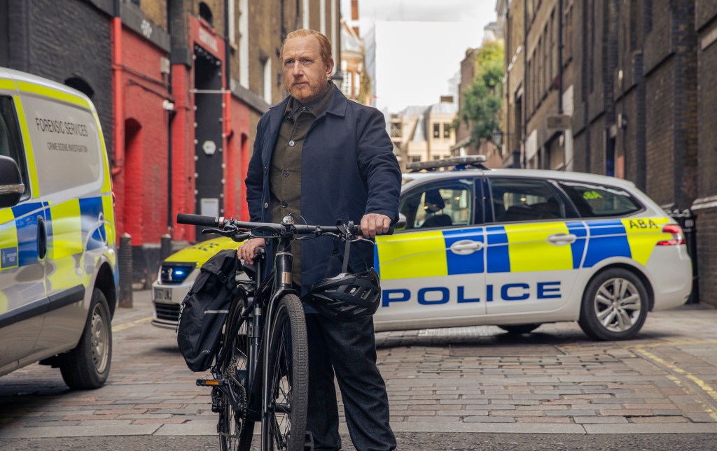 Adrian Scarborough as DI Max Arnold. He's at a crime scene and is holding his bicycle. There are two police cars behind him.