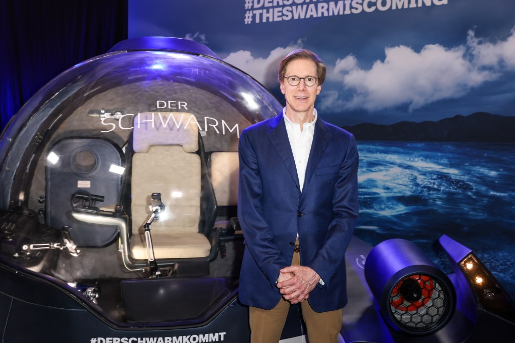 Photo of series executive producer Frank Doelger at the premiere of "The Swarm" in Germany. He's standing in front of an undersea robotic vessel and is wearing a blue sports jacket with a white shirt and brown pants. He's got his hands crossed in front of him.