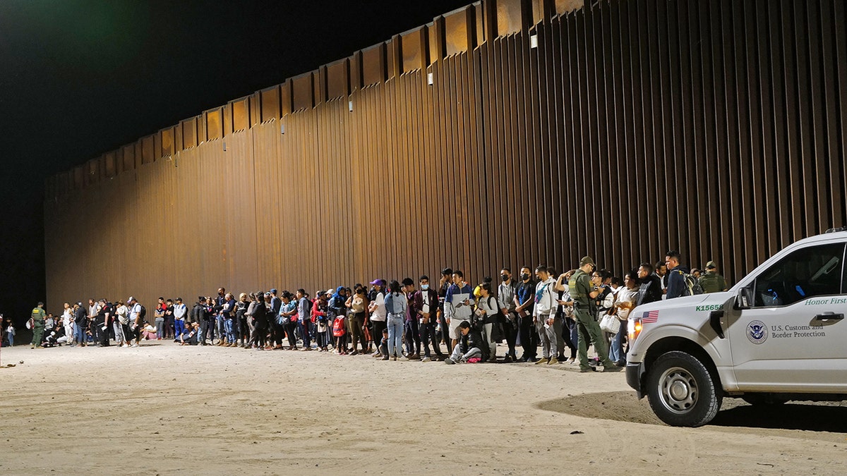 Migrants standing in a line along the border wall