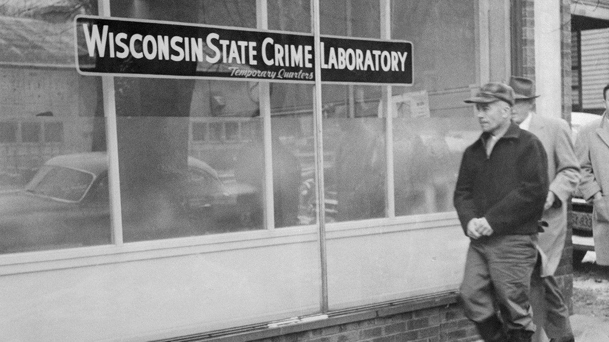 Ed Gein in a dark jacket and hat walking towards a building