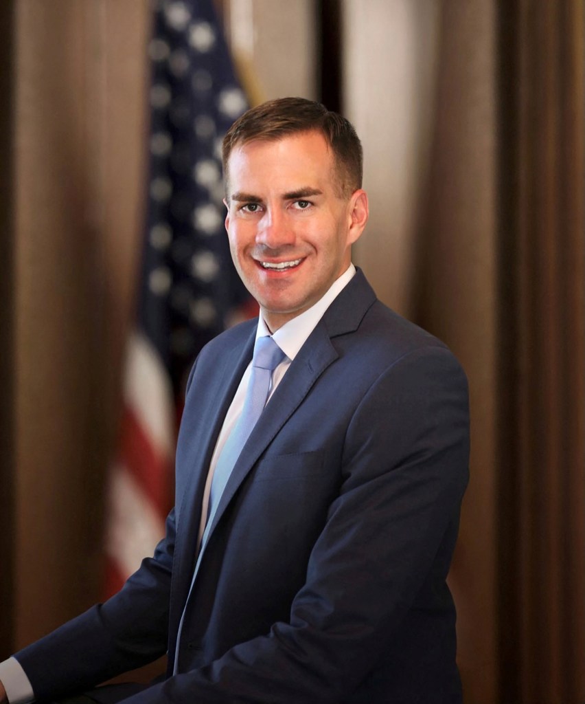 Colin Schmitt in coat and tie in front of a blurry American flag. 