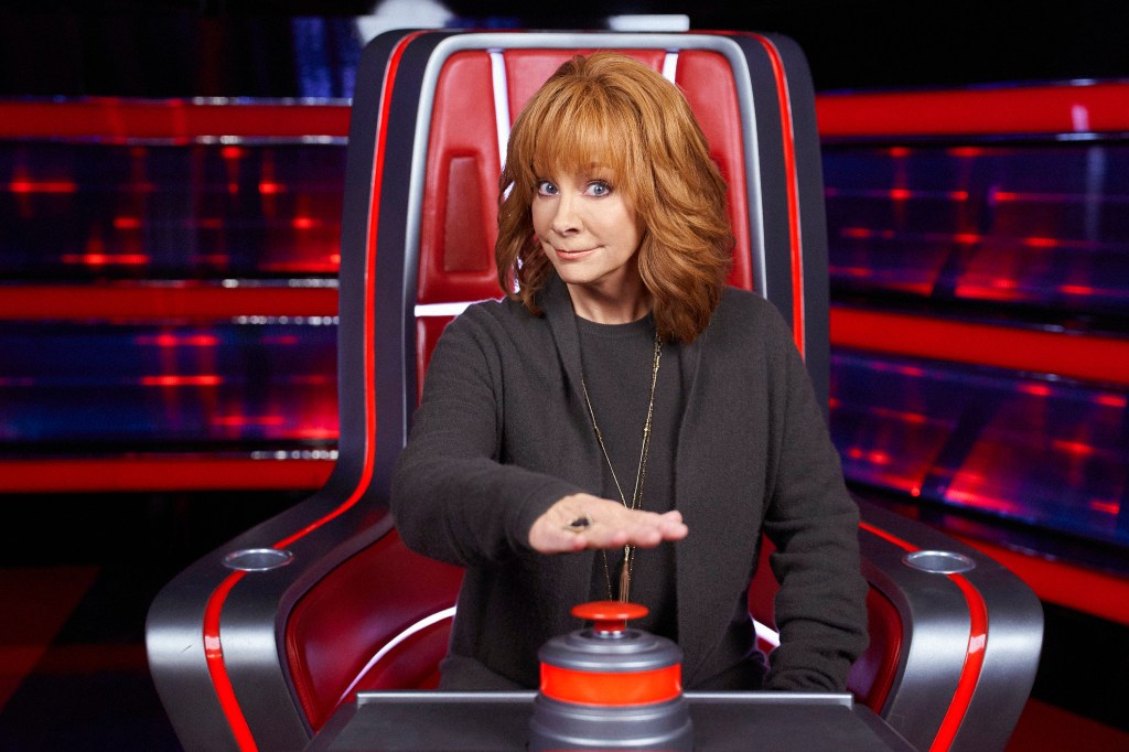 Reba McEntire sitting in a chair in front of a red button. 