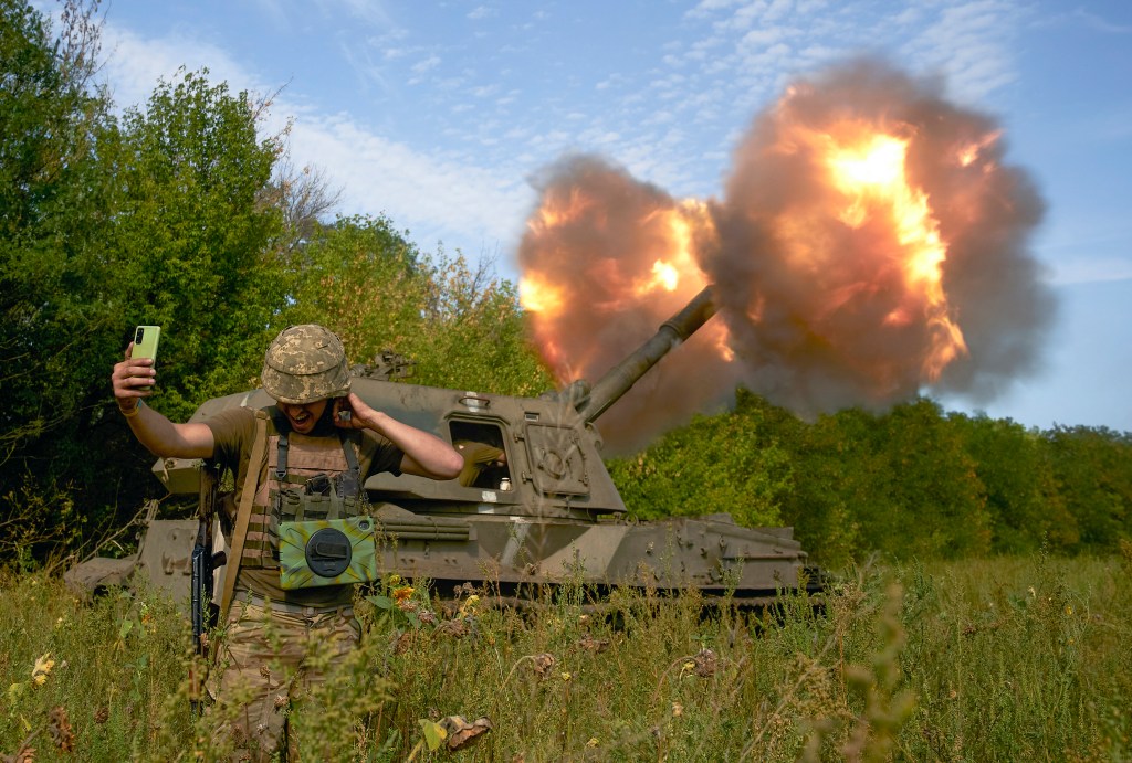 A Ukranian soldier takes a selfie in front of an artillery vehicle as it fires.