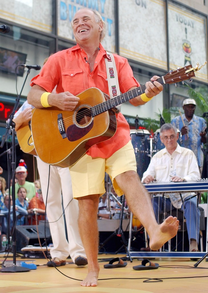 A photo of Jimmy Buffett barefoot and holding a guitar on stage.