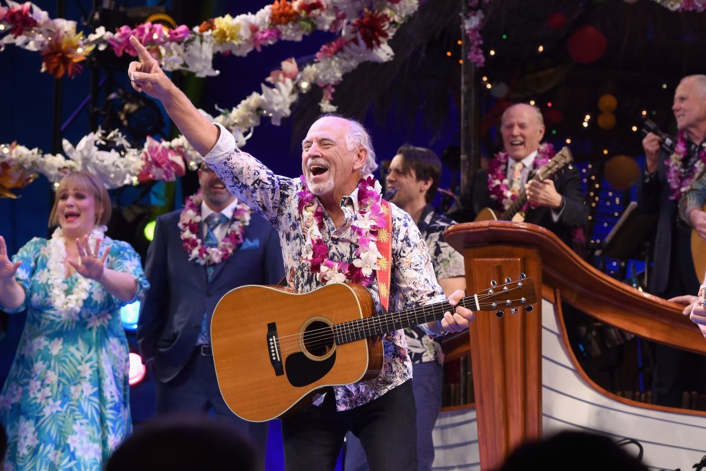 Singer immy Buffett smiles and gestures while holding a guitar on a stage decorated with flowers.