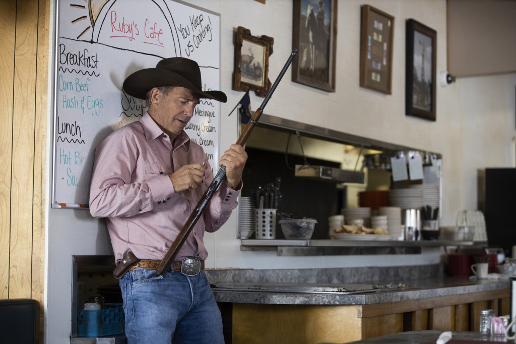 Kevin Costner holding a rifle in a kitchen wearing a cowboy hat. 