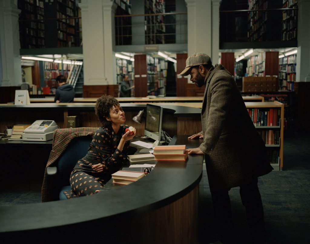 Emma (Clark Backo) and Apollo (LaKeith Stanfield) flirting in the library. She sits behind a desk, he stands. 