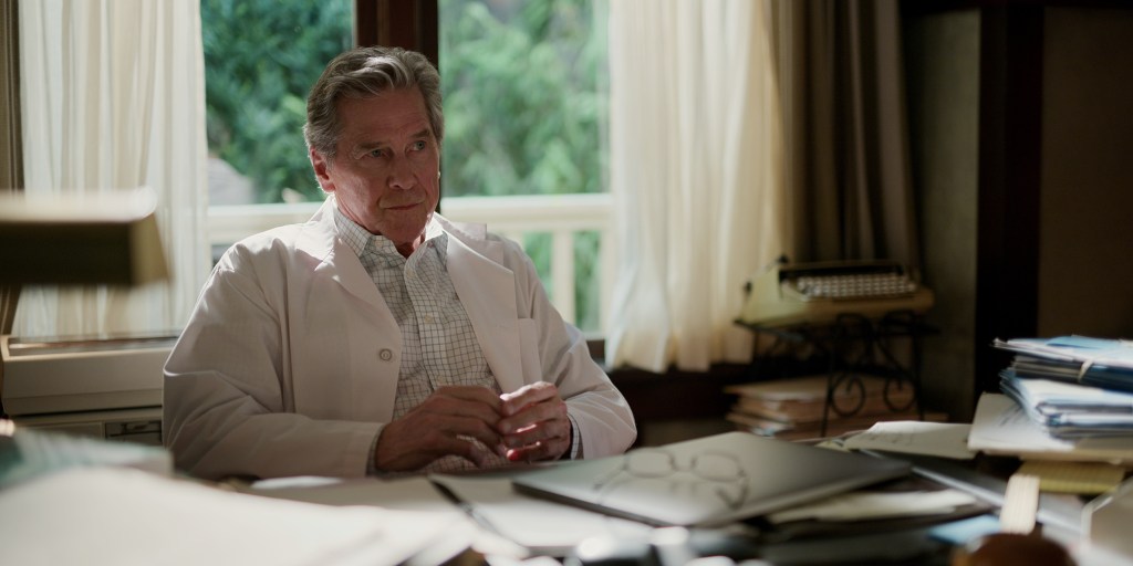 Doc Mullins (Tim Matheson) sits in his office chair. He's staring straight ahead and is wearing a white doctor's jacket.