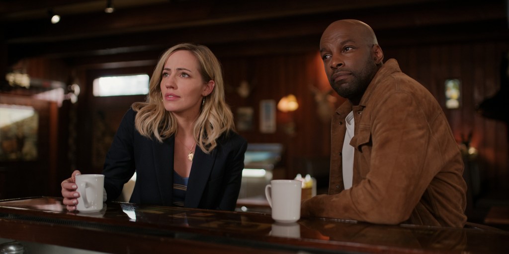 Brie (Zibby Allen) and Preacher (Colin Lawrence). They're sitting in a bar with cups of coffee in front of them. They're looking off-camera at something.