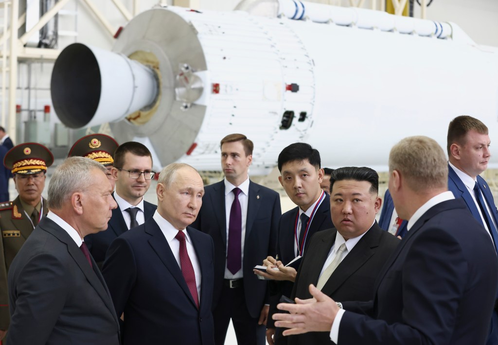 Russian President Vladimir Putin and North Korean leader Kim Jong Un listen to a speaker with his back to the camera, with aides surrounding them, at a rocket assembly plant in Russia.
