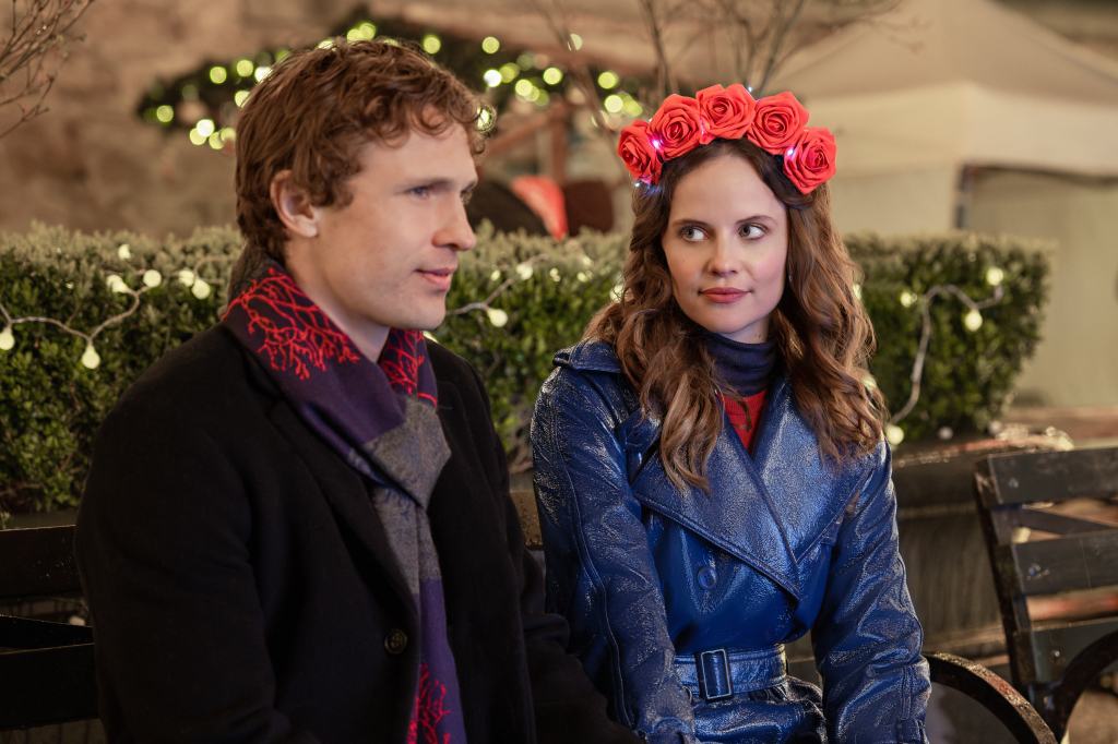William Moseley and Sarah Ramos in "Christmas in Notting Hill" sit in front of a christmas tree, smiling at each other. 