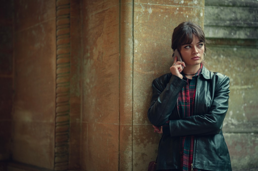 Emma Mackey leaning against a wall holding a phone looking serious. 
