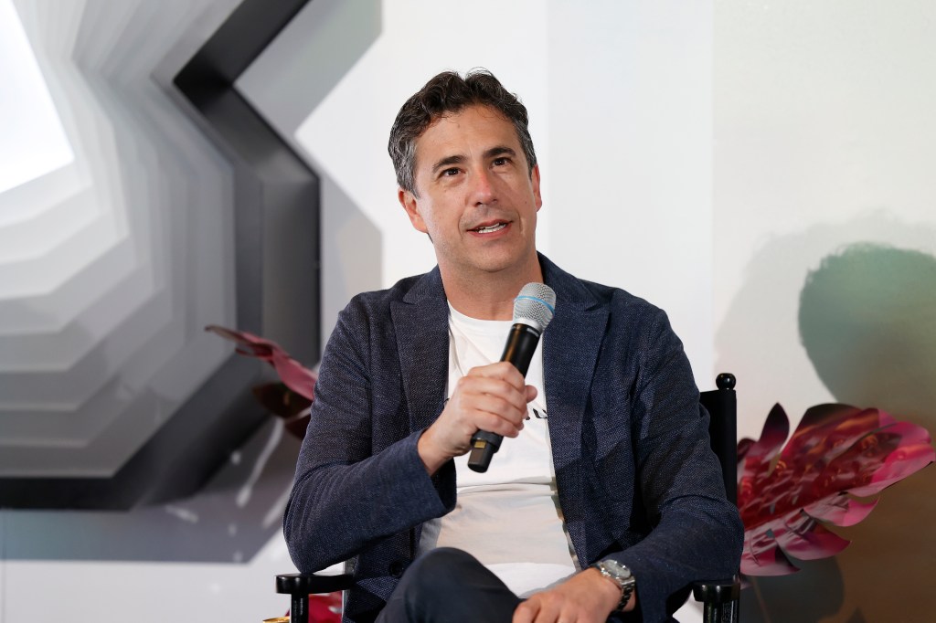 Michael Thorn is sitting in a director's chair on a stage and is holding a microphone. He's dressed casually in a blue jacket and white T-shirt.