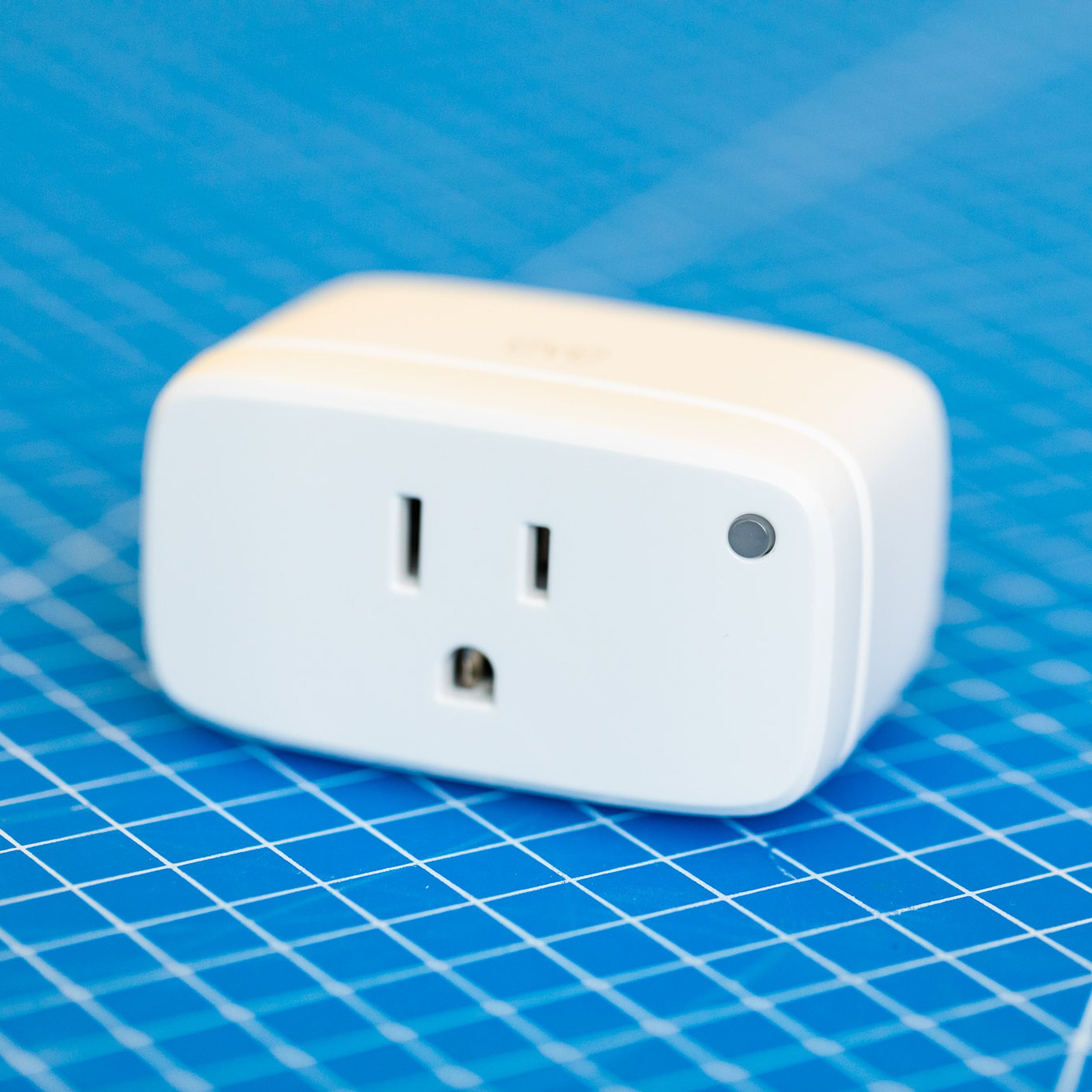 Eve Energy smart plug. A white rectangular prism with the broad face front. The corners and edges are rounded, there is a three-prong outlet on the front and a green LED on the upper-right corner of the front face. It sits on a blue background with a grid of white lines.