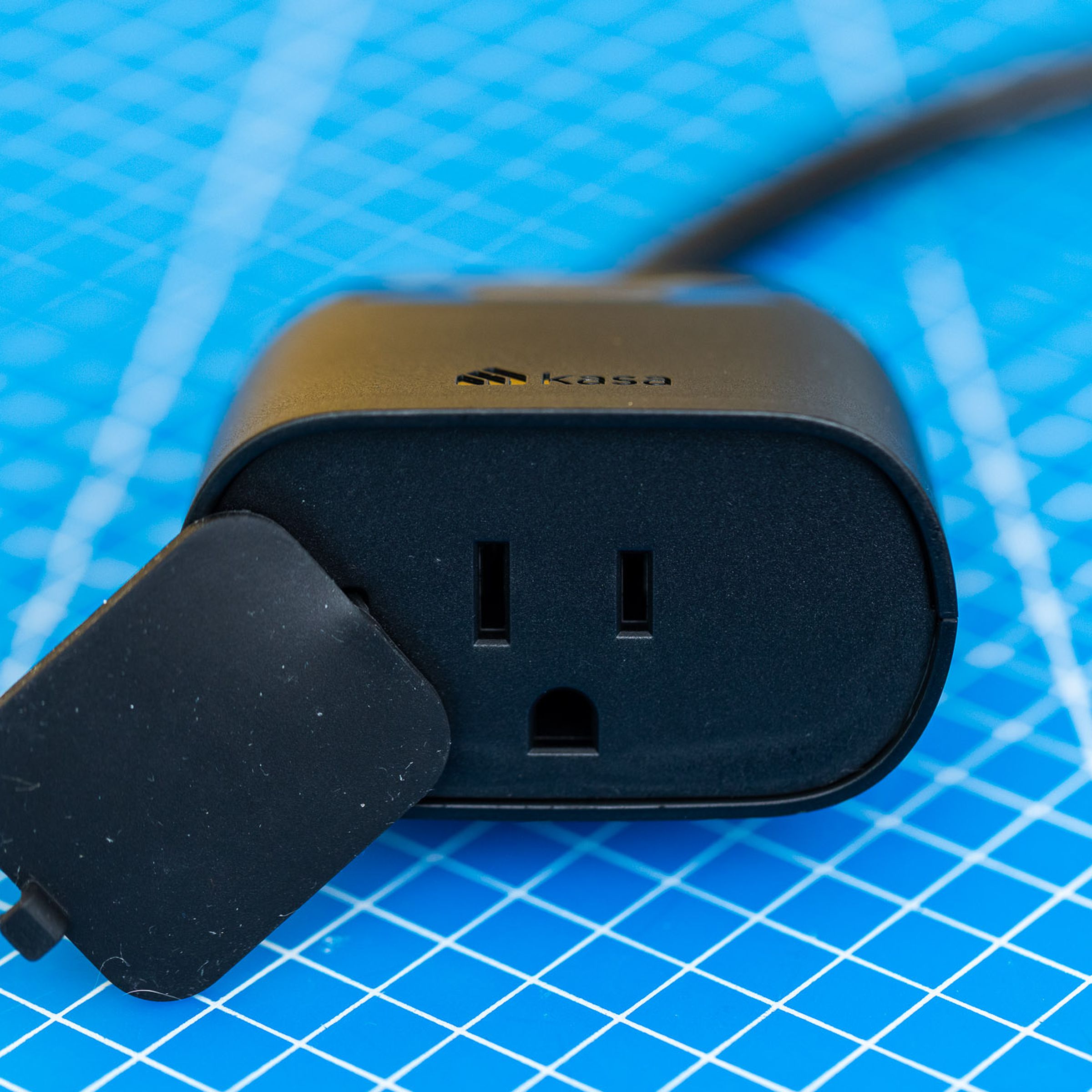 A black, two-outlet outdoor smart plug. It has a short cable leading to its own power plug, and a flap to protect the outlets from moisture. It is on a blue background with a grid of white lines.