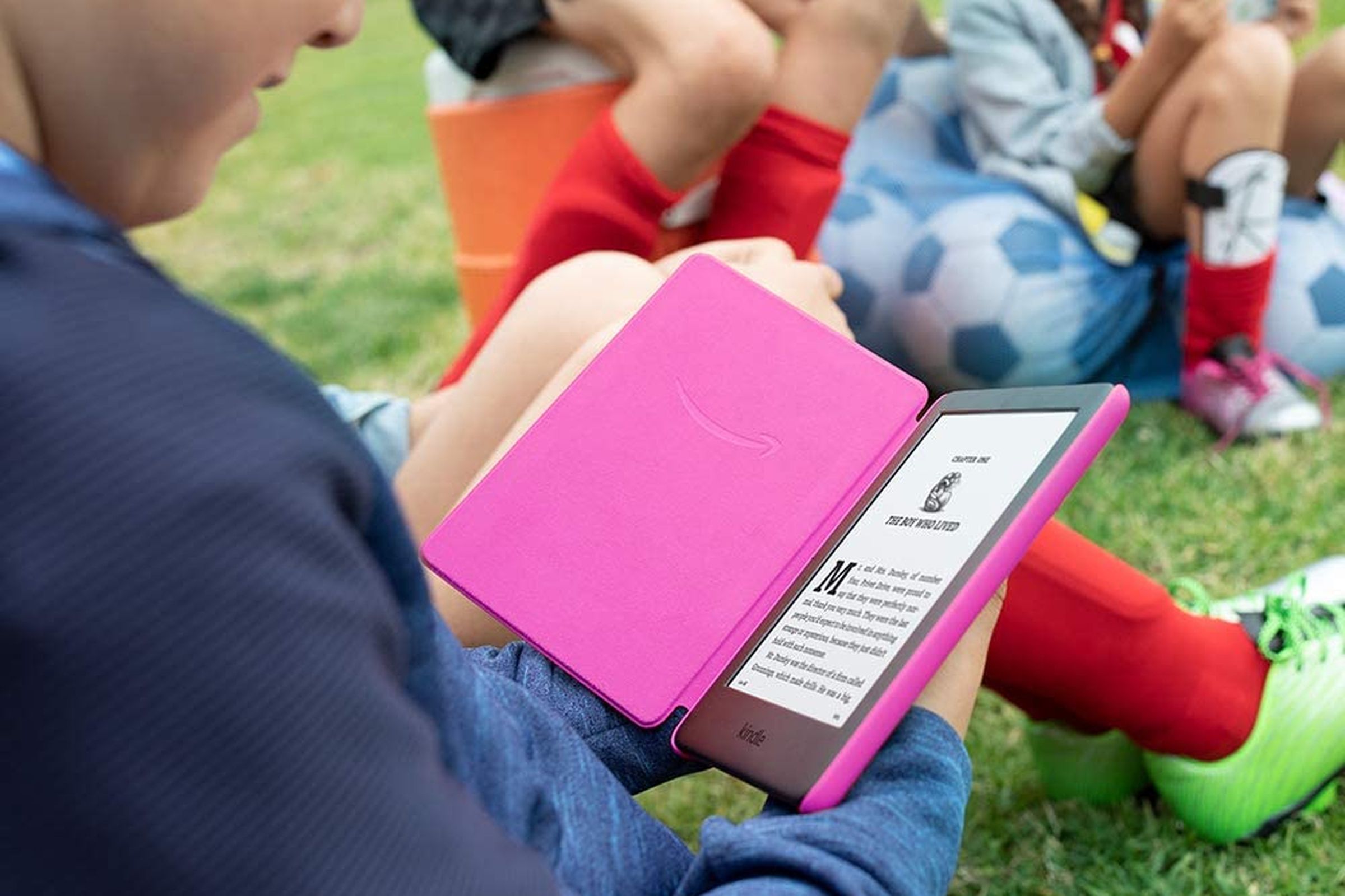 A child holding and reading a pink Kindle while sitting on grass.