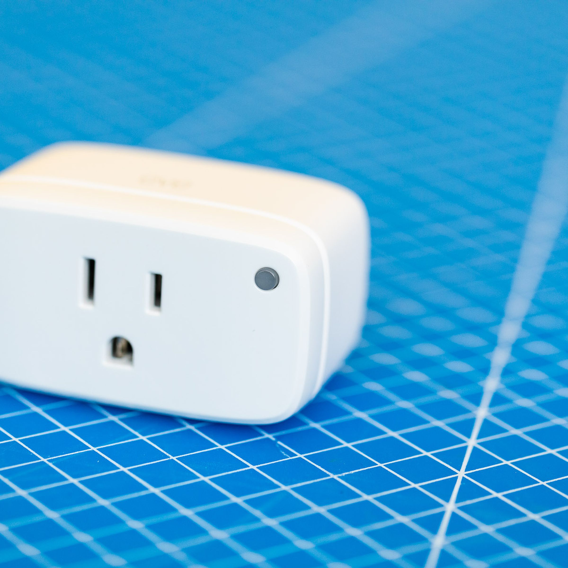 Eve Energy smart plug. A white rectangular prism with the broad face front. The corners and edges are rounded, there is a three-prong outlet on the front and a green LED on the upper-right corner of the front face. It sits on a blue background with a grid of white lines.