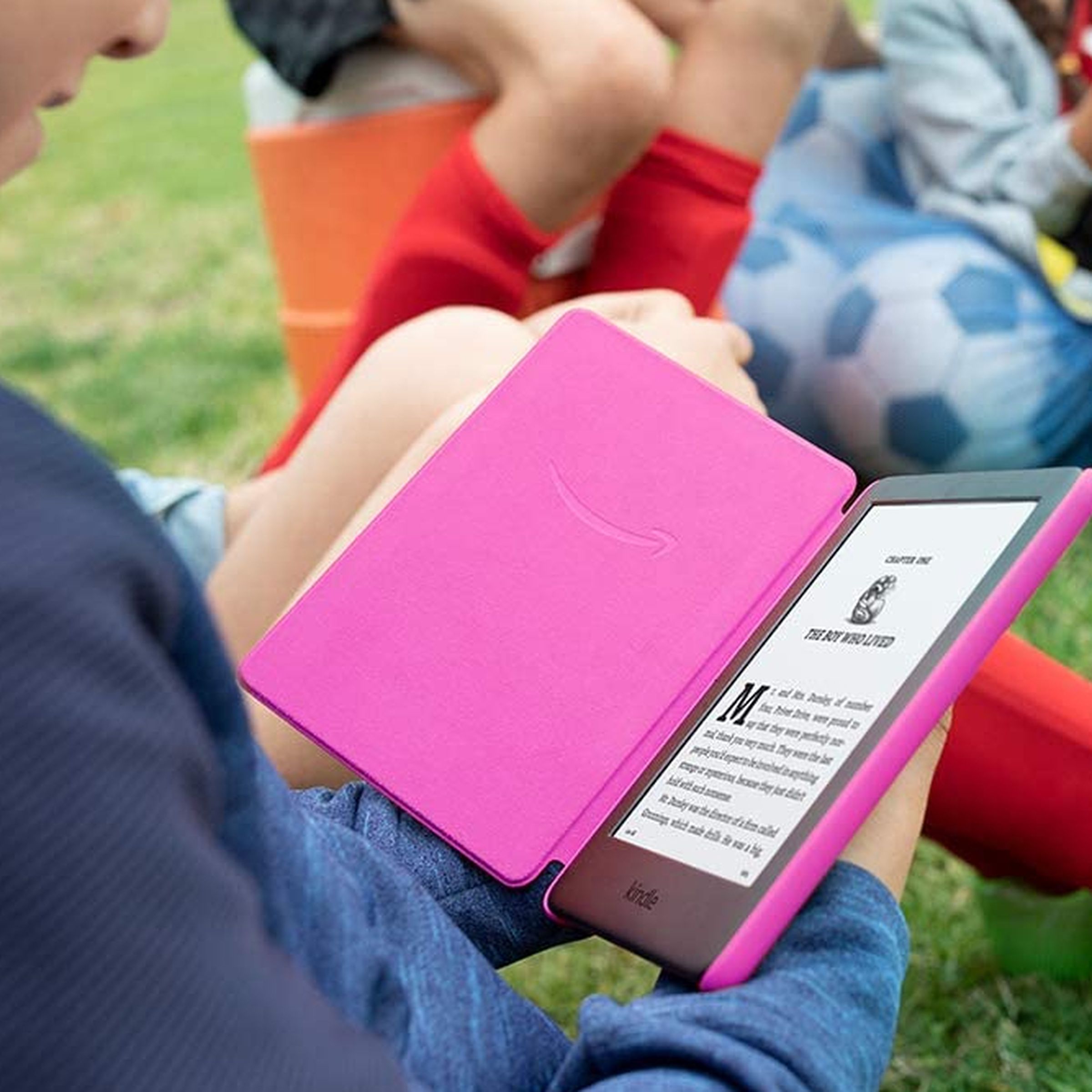 A child holding and reading a pink Kindle while sitting on grass.