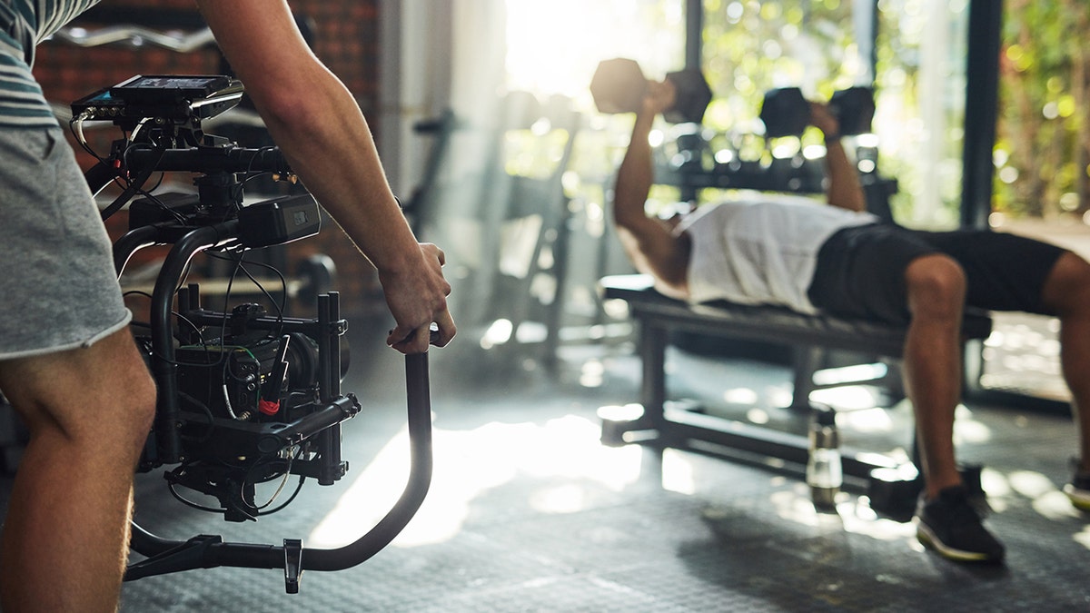 Person films man lifting weights while lying on a bench at a gym