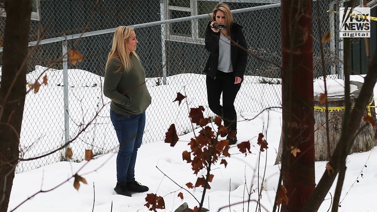 Two women stand in the snow at the back of a house while one of them uses a video camera.