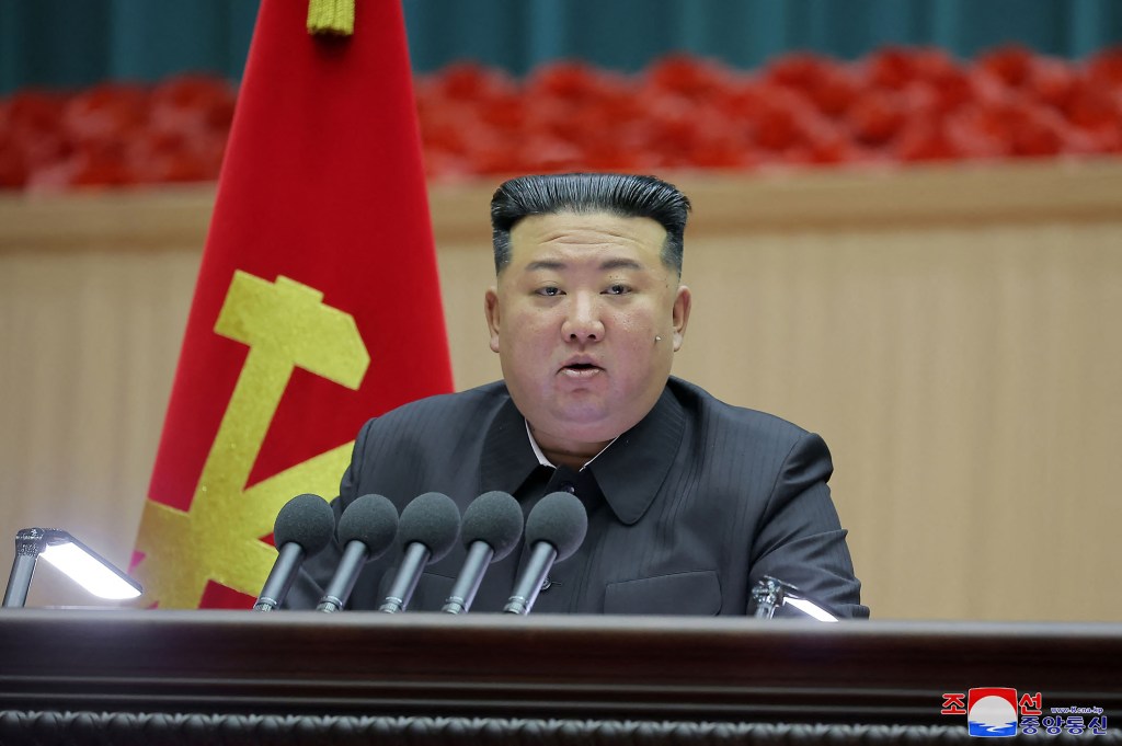 North Korean leader Kim John Un sits in front of a bank of five microphones with the North Korean flag on a pole behind his right shoulder.
