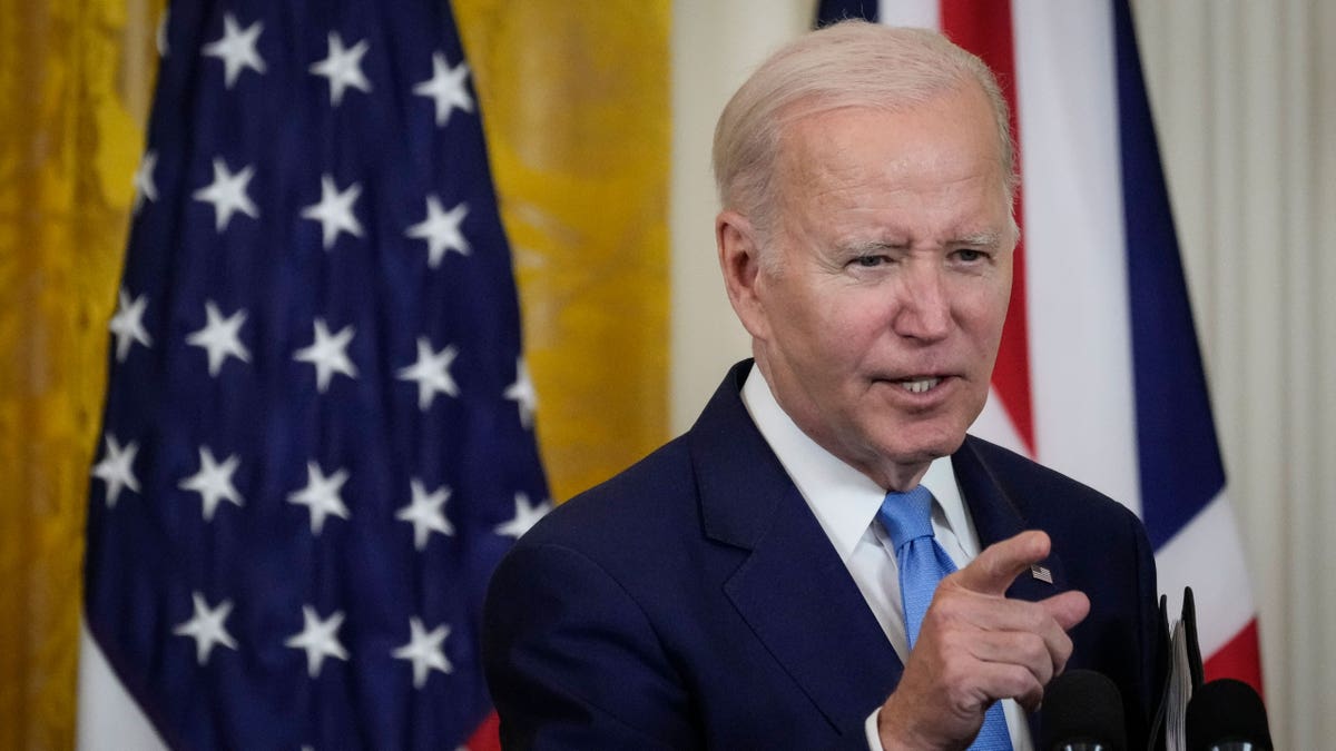 President Joe Biden gesturing with right hand, US, UK flags behind him