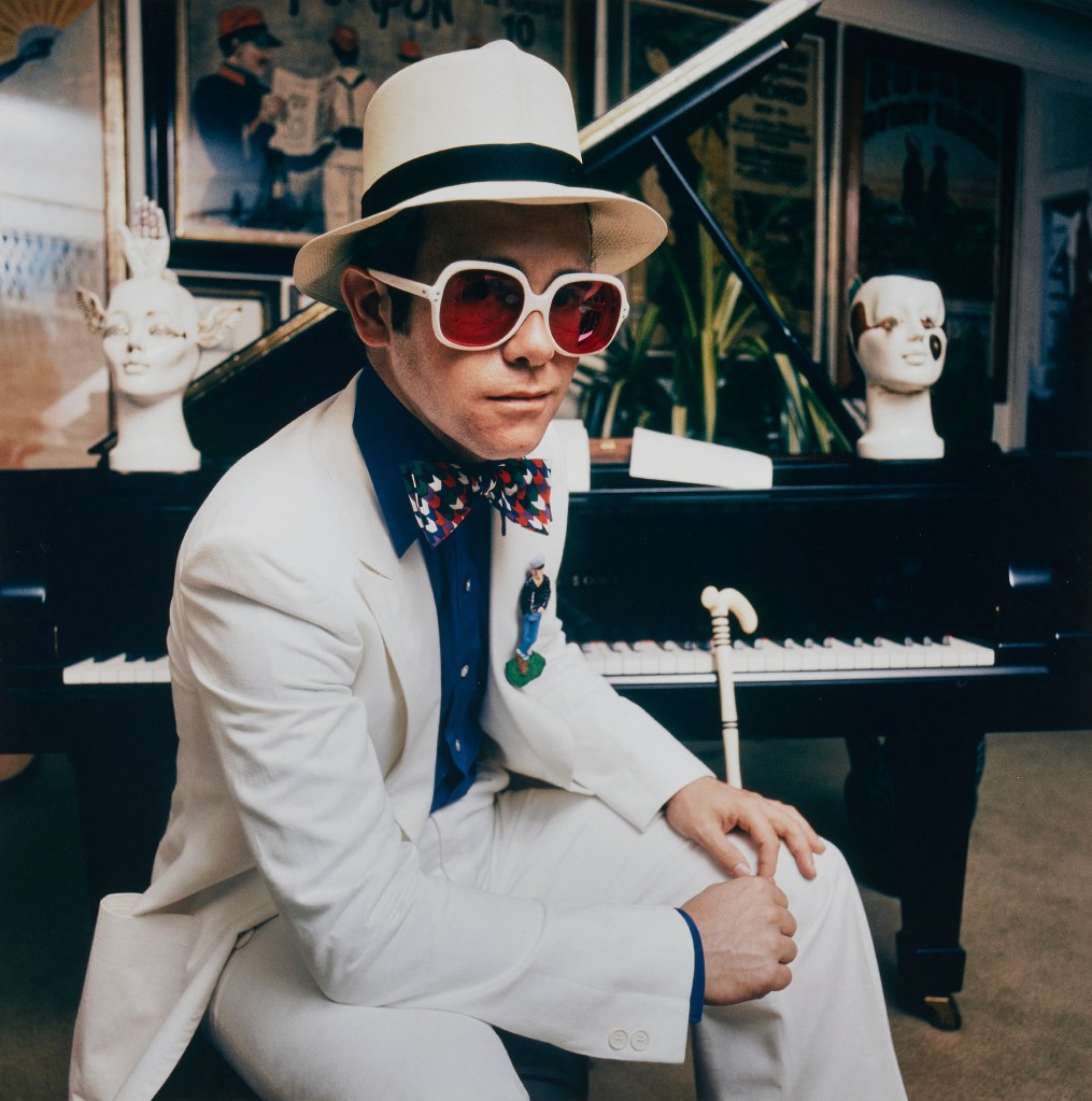 Elton John wearing a white suite, hat and large white-framed sunglasses, sitting at a grand piano.