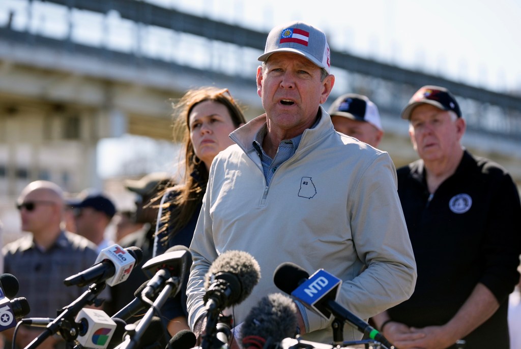 Georgia Gov. Brian Kemp speaking at a news conference about border policies, surrounded by microphones and press in Shelby Park.