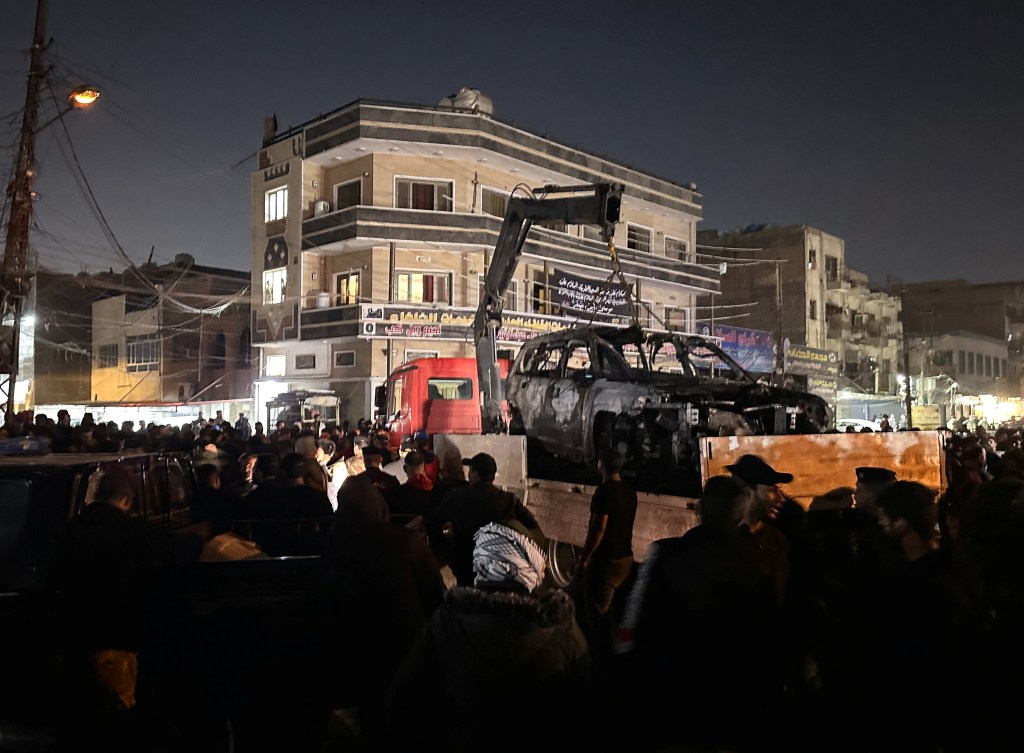 Destroyed vehicle on a loaded truck in Baghdad after a deadly drone strike: people gather around it.